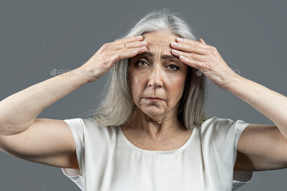 Sad old european lady with gray hair touches her face, wrinkles with hands, isolated on gray