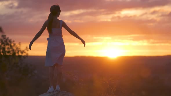 Dark Silhouette of a Young Woman Standing with Raised Up Hands on a Stone Enjoying Sunset View alt