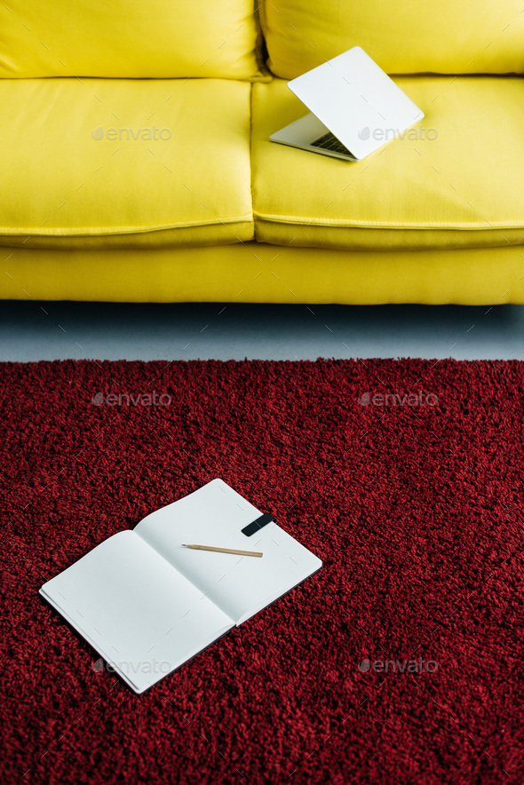 Half-closed laptop on yellow couch and textbook with pencil on rug