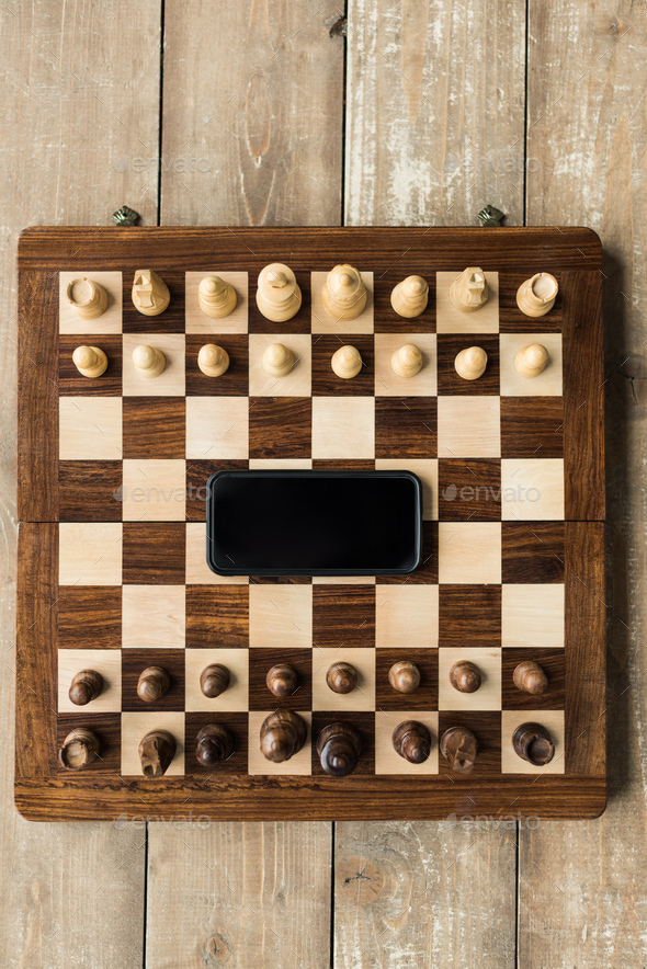 Top view of chess board with smartphone and chess pieces on wooden surface