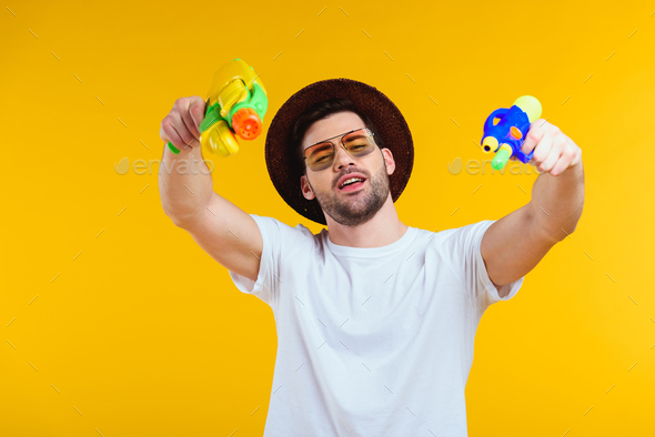 stylish young man in hat and sunglasses holding water guns and looking at camera isolated on yellow
