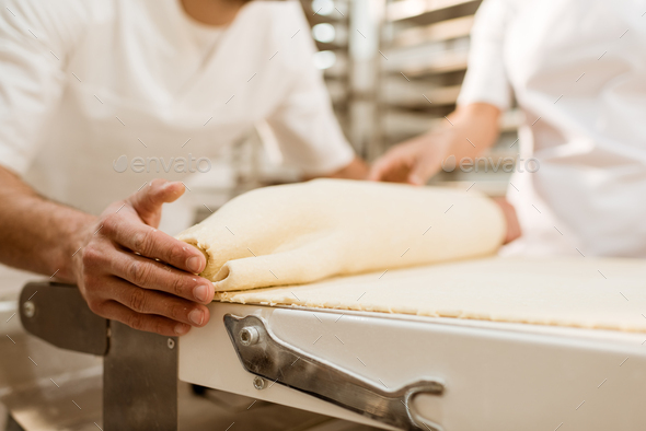 cropped shot of bakers rolling dough with industrial dough roller at baking manufacture