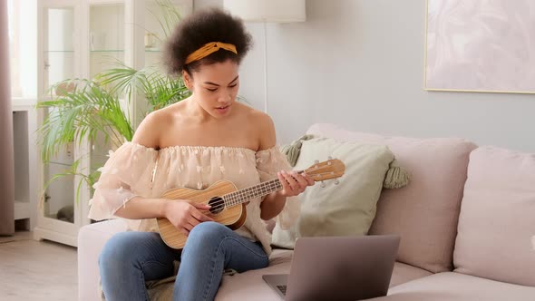 Young african american woman taking online ukulele music guitar lessons tutorial on laptop alt