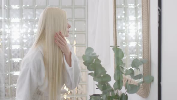 Side View of Young Caucasian Woman With Long Blond Hair Wearing a Bath Towel Looking in the Mirror alt