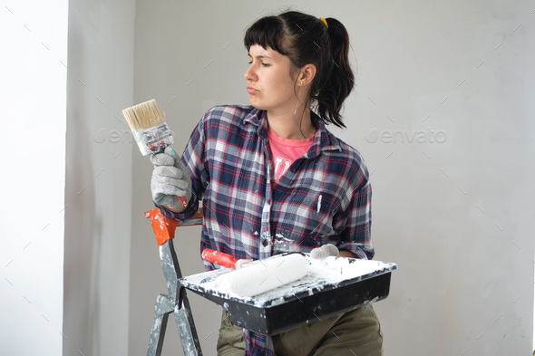 Woman in paint roller and white paint paints the wall of house on stepladder