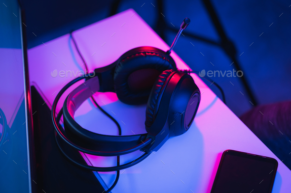 High angle shot of gaming headphones and a phone on a desk under colorful led lights in a dark room