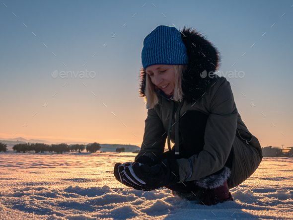 Woman crouching down and collecting snow in a beautiful snow field during the sunset