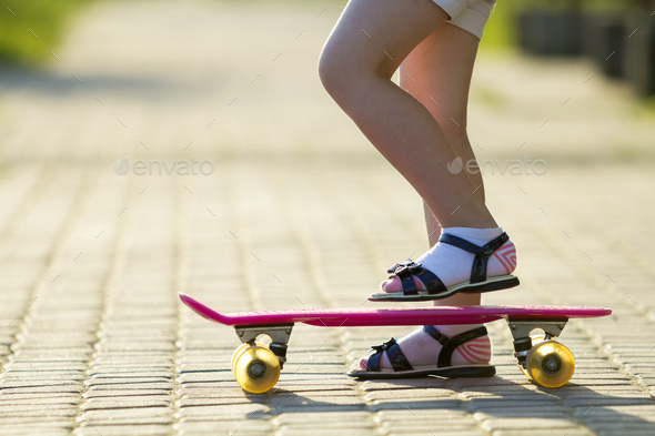 Child slim legs in white socks and black sandals on plastic pink skateboard