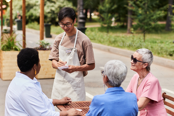 Young Asian waiter serving group of senior women at outdoor cafe