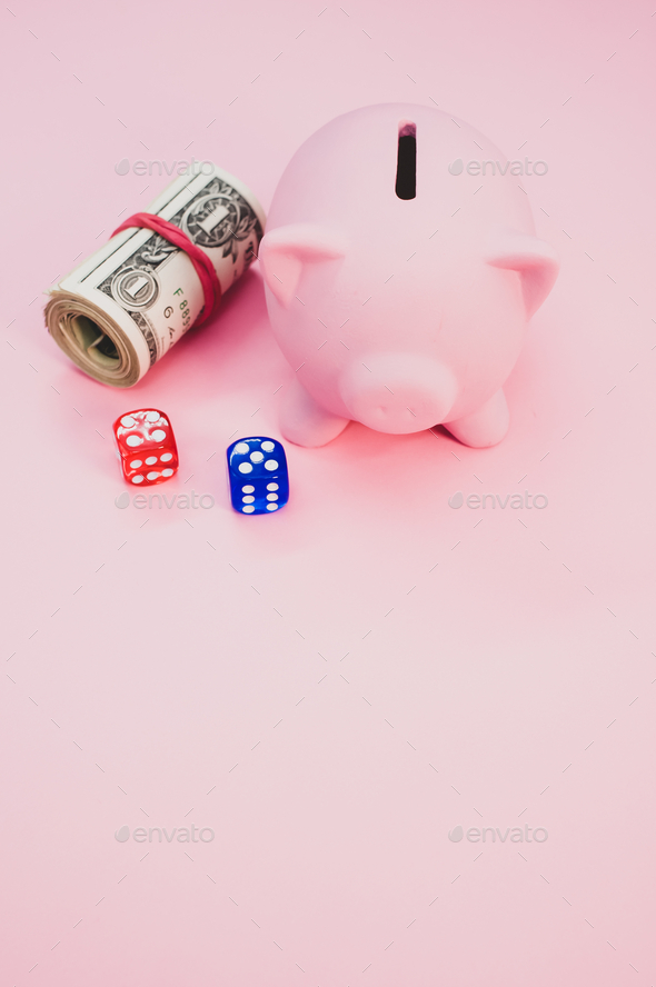 Vertical shot of a piggy bank next to a roll of dollars and dice isolated  on pink background