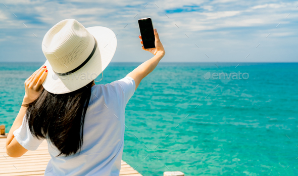 Young Asian woman wear straw hat in casual style use smartphone taking selfie at wooden pier. Summer - Stock Photo - Images