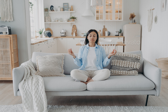 Female practices yoga, breathing, relaxing on cozy couch at home. Healthy lifestyle, stress relief