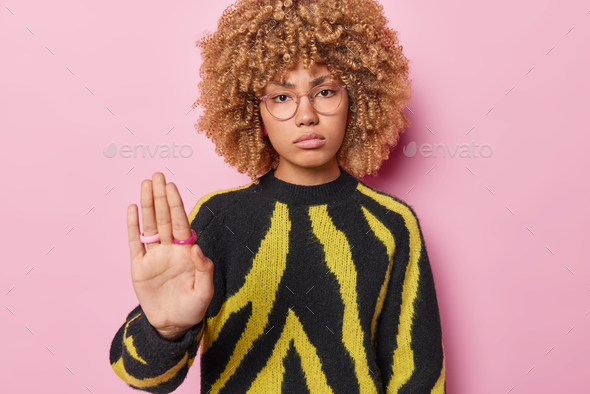 Portrait of serious curly haired European woman makes stop gesture keeps palm raised towards camera