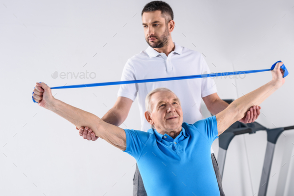 portrait of rehabilitation therapist assisting senior man exercising with rubber tape