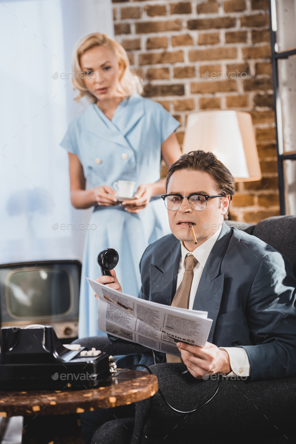angry man with cigarette holding newspaper and vintage telephone while woman holding cup of coffee