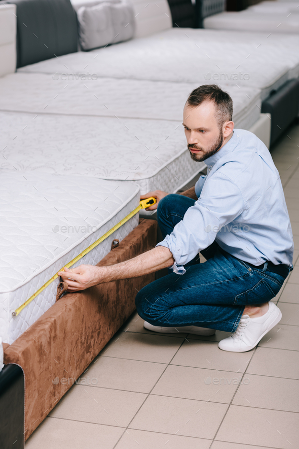 male shopper measuring orthopedic mattress with measure tape in furniture store