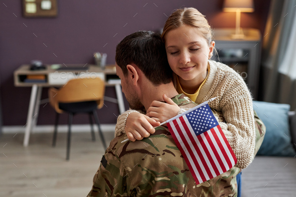 Teen girl embracing military dad and holding american flag at home
