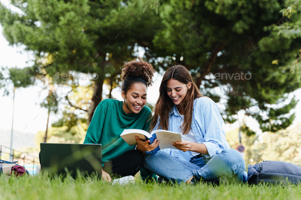 Women students sitting on public park grass learning together with book and laptop