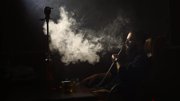 Young bearded man smokes a hookah and blow out smoke on black background in slowmotion