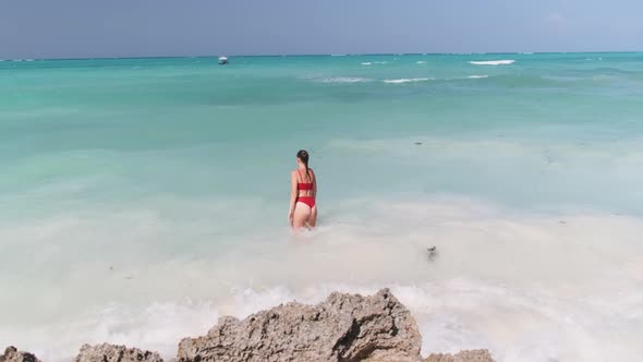 Young Woman in a Red Swimsuit Walks Into the Turquoise Ocean on a Paradise Beach alt