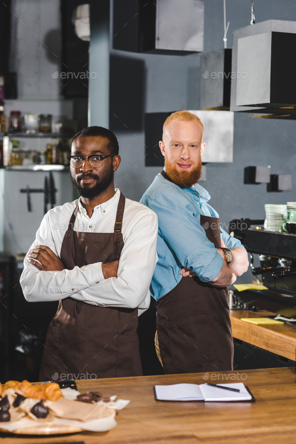 two young multicultural male baristas in aprons standing with crossed