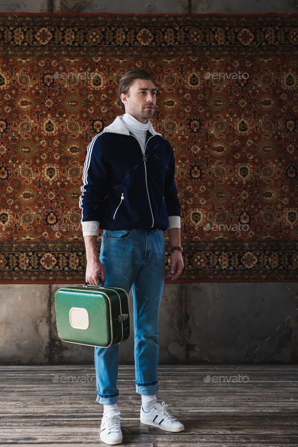 handsome man with vintage little suitcase in front of rug hanging on wall