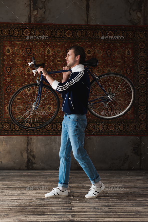 handsome young man in vintage clothes carrying road bike on shoulder in front of rug hanging on wall