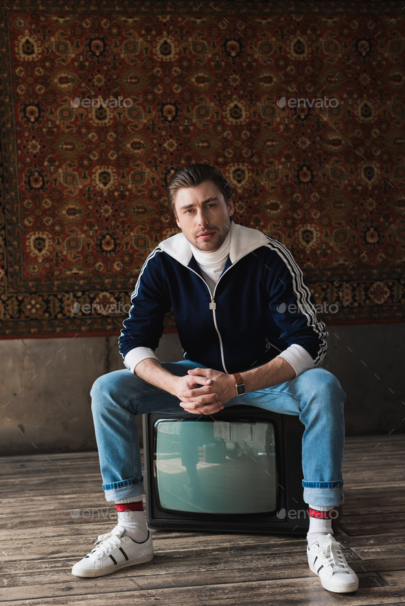 handsome young man in vintage clothes sitting on retro tv set and looking at camera in front of rug