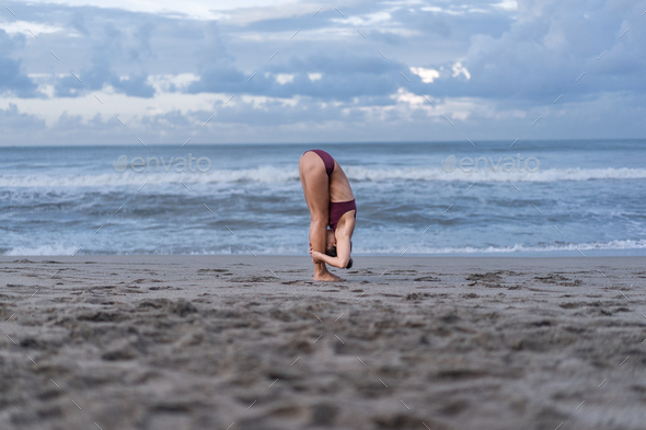 side view of young woman practicing yoga in Standing Forward Bend pose (Uttanasana) on seashore