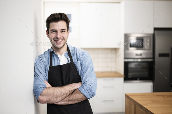 handsome young man smiling in the kitchen wearing an apron - Portrait of a cheerful confident - Stock Photo - Images