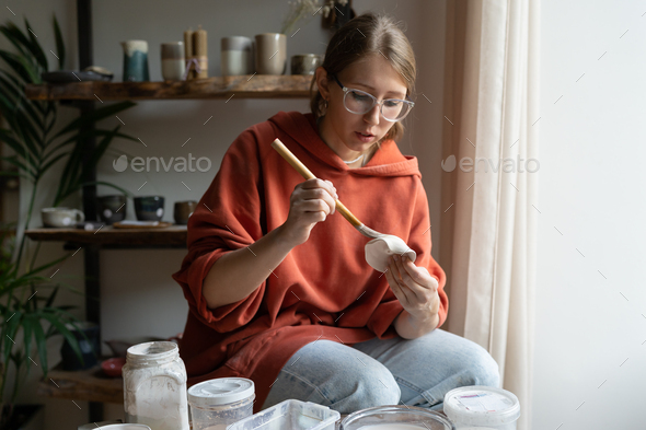 Creative focused girl artist uses brush to draw pattern on clay products during pre-sale preparation