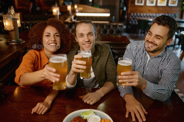 Cheerful friends drinking draft beer at bar table in pub