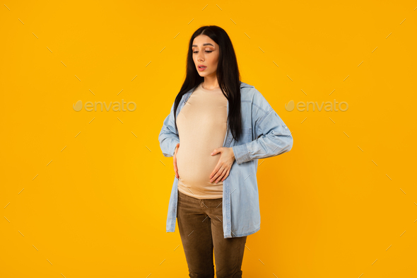 Pregnant lady having painful spasm and touching belly, doing breathing exercises over yellow