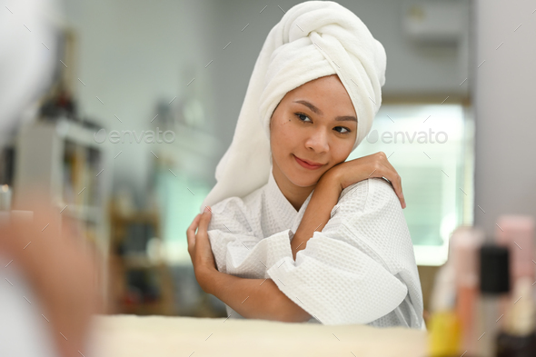 Beautiful young woman with towel on head hugging herself and looking in mirror.