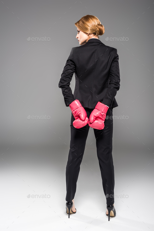 rear view of businesswoman posing in suit and pink boxing gloves, isolated on grey
