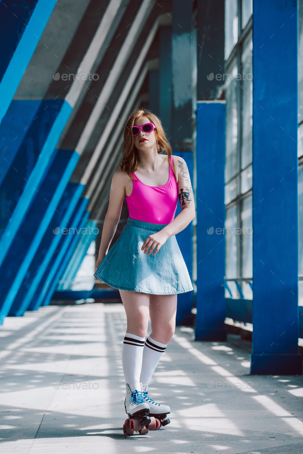 beautiful stylish girl in pink sunglasses and denim skirt roller skating and looking at camera