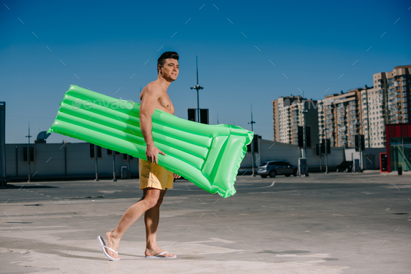 side view of young shirtless man with inflatable bed walking by parking