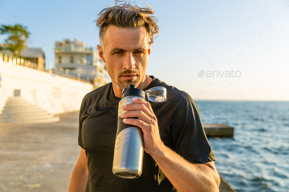 adult sportsman drinking water from fitness bottle on seashore in front ...