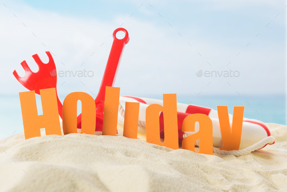 Beach toys and Holiday inscription in sand on blue sky background