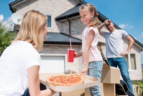 side view of happy mother holding box with pizza and daughter with cup of cola taking slice of pizza