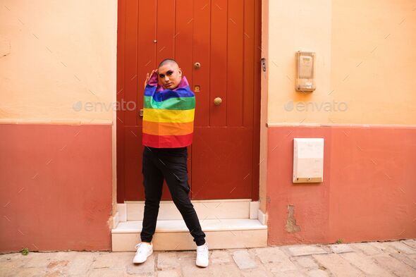Hispanic boy with vibrant makeup carrying gay pride flag on his shoulders against red wooden door