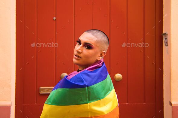 Hispanic boy with vibrant makeup carrying gay pride flag on his shoulders against red wooden door