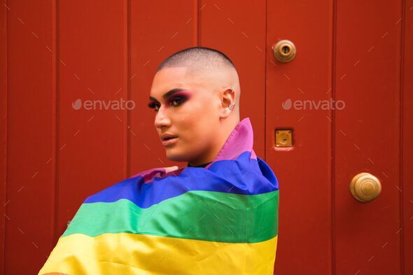 Hispanic boy with vibrant makeup carrying gay pride flag o his shoulders against red wooden door