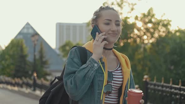 Joyful Hipster Girl Using Cellphone Outdoors with Cup of Coffee or Tea in Hand alt