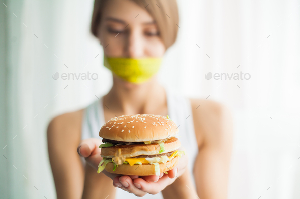 Young woman with duct tape over her mouth, preventing her to eat junk food. Healthy eating concept
