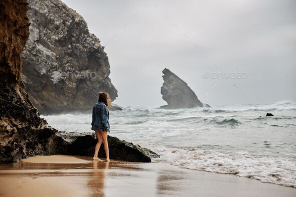 Female in a jeans coat walking on the wet sand washed by the waters of the turbulent sea