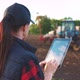 Woman Uses Specialized App on a Digital Tablet PC on Background of Working Tractor in Field - VideoHive Item for Sale