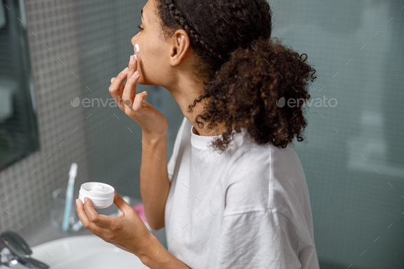 African Woman applying moisturizing cream standing behind mirror in home bathroom. Beauty procedure