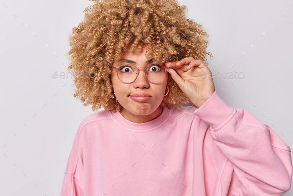 Close up shot of curly haired adult woman stares amazed at camera keeps hand on rim of spectacles