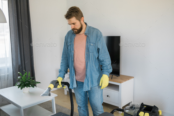 Man holding modern washing vacuum cleaner and cleaning dirty sofa with professionally detergent